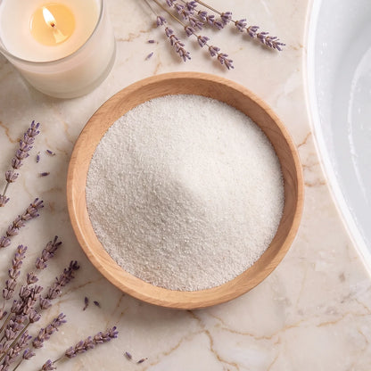 Top-down view of pure white Epsom Salt crystals in a wooden bowl, surrounded by fresh lavender sprigs and a lit candle on marble.