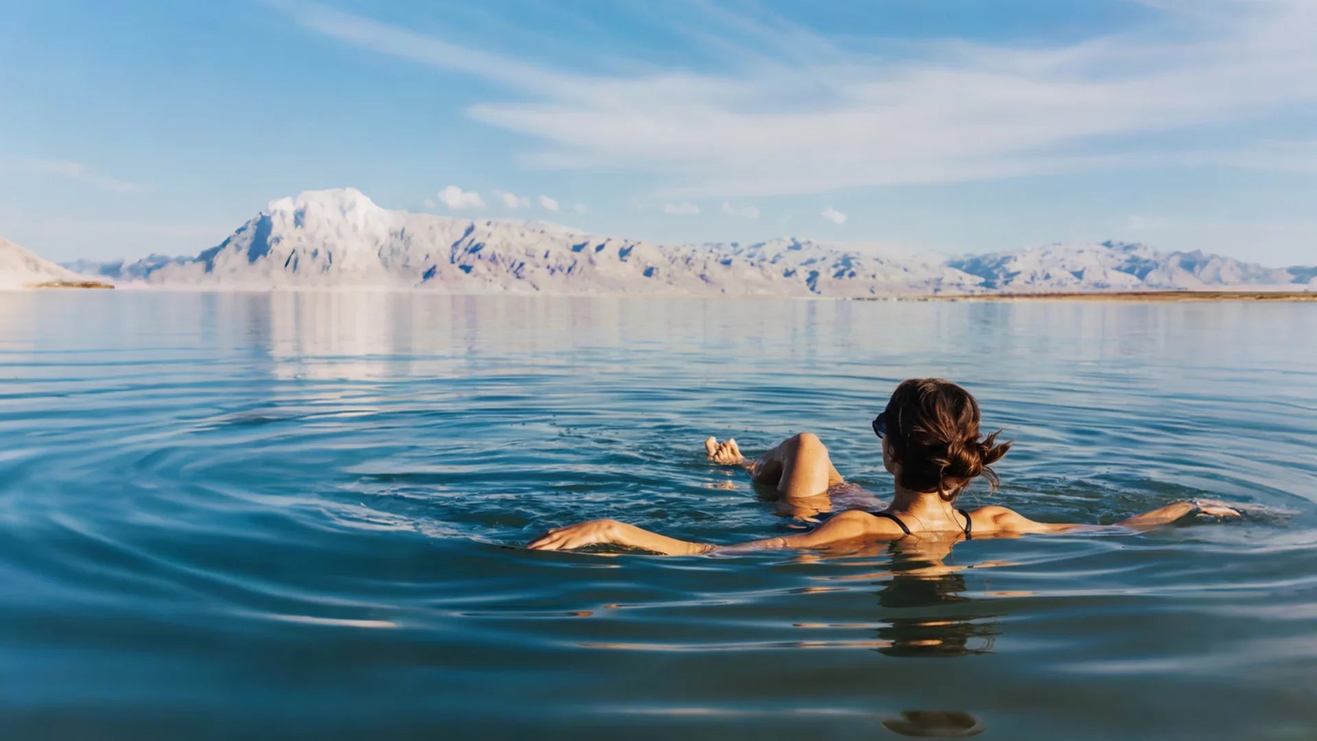 A woman floats peacefully on a calm blue lake with snow-capped mountains in the distance, embodying the deep relaxation from a soothing Epsom Salt soak.